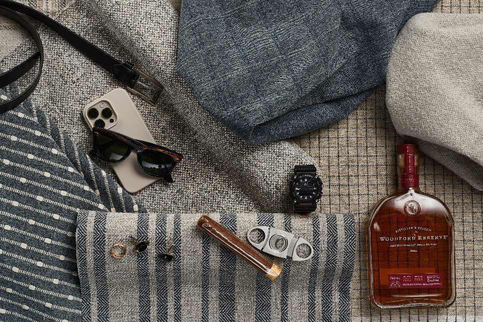 Spread of mens clothing, belt, phone, sunglasses, watch, cigar, whiskey, and cufflinks.